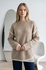 Sweater de tejido de punto en color beige con textura suave y detalles de costuras visibles en color blanco en los hombros, puños y ruedo. Presenta un diseño de corte holgado, cuello redondo y mangas largas.