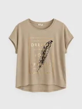 Remera de algodón color beige con corte relajado y diseño asimétrico, más larga en la parte posterior. Presenta un estampado frontal rectangular con letras doradas, una pluma ilustrada y detalles de pedrería decorativa.