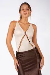 Body de color beige con escote en V profundo y diseño frontal con aberturas unidas por argollas metálicas doradas. Presenta breteles anchos y un ajuste al cuerpo.