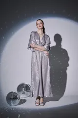 Vestido largo de fiesta en color plateado con acabado brillante. Presenta escote en V cruzado, mangas tipo kimono 3/4 y cintura elástica con frunces que dan caída fluida a la falda.