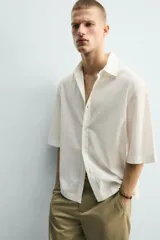 Camisa de corte relajado y boxy fit, confeccionada en tejido con textura irregular. Presenta cuello solapa, manga corta y cierre frontal con botones.