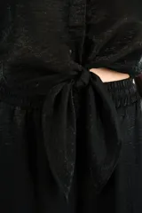 Camisa crop negra de tejido fluido y acabado brillante, con cuello camisero, mangas cortas con vuelta y nudo frontal en el dobladillo.