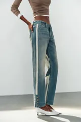 Jeans rectos de tiro medio en denim azul con efecto deslavado. Presentan bandas laterales con detalles brillantes, bolsillos delanteros y traseros, y aberturas en los tobillos con botonadura metálica.