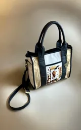 Cartera de cuero modelo Tali, de diseño estructurado que combina los colores blanco, negro y dorado, con detalles de arte pintado a mano en el frente. Cuenta con doble asa de mano, correa larga desmontable y herrajes metálicos.