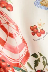 Vestido mini de color beige con un estampado de tomates, aceitunas y botellas de aceite, y un borde inferior rojo con ilustraciones de una sombrilla de playa y vegetales. Cuenta con escote cuadrado y tirantes anchos cruzados en la espalda.