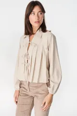 Blusa de algodón color beige con cuello camisero, mangas largas abullonadas con puños elásticos y cierre frontal mediante tres lazos para anudar.