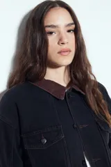 Campera de jean color negro con cuello de pana en tono marrón, cierre frontal con botones y bolsillos en el pecho. Presenta un diseño de corte relajado y ligeramente cropped, con interior forrado en corderito para mayor abrigo.