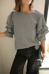 Blusa de tela con estampado vichy en blanco y negro, con cuello redondo y mangas con tres volados escalonados que llegan hasta el codo. Presenta puños elásticos y cierre con botón en la parte posterior.