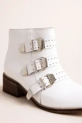 Botineta blanca de cuero, con taco cuadrado de madera, punta afilada y tres tiras con hebillas metálicas y tachas decorativas.