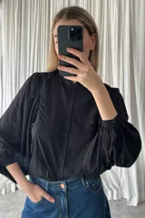 Camisa negra de corte clásico con cuello alto tipo mao con volados, cierre frontal de botones y mangas largas abullonadas con puños ajustados.