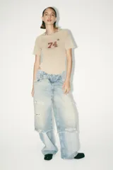 Camiseta color beige de algodón con efecto lavado, corte regular, cuello redondo con tachas metálicas, mangas cortas y estampado frontal con el número 74 y una estrella.