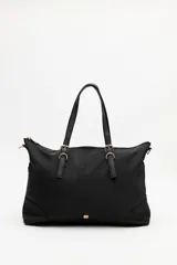Cartera tipo tote de nylon negro, con diseño amplio y liviano. Presenta asas superiores fijas con detalles de tiras regulables y herrajes metálicos dorados. Incluye cierre principal y un pequeño logo metálico en la parte inferior frontal.