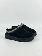 Zueco de gamuza negra con interior forrado en corderito sintético. Presenta un detalle de bordado geométrico en el borde superior y una suela de plataforma plana.