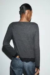 Top de manga larga en tejido suave color gris, con cuello redondo y bajo asimétrico decorado con encaje negro a tono. Presenta detalles de encaje negro en los puños.