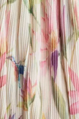 Vestido corto con textura plisada, estampado floral en tonos pastel (rosa, verde, amarillo, azul) sobre fondo crema. Presenta escote pronunciado en V con fruncido central y mangas globo largas ajustadas con lazos en los puños.