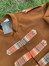Chaqueta de estilo militar en color marrón, confeccionada en tejido de lana. Presenta un diseño estructurado con cuello mao y detalles decorativos en los laterales frontales con aplicaciones de tela a cuadros y botones metálicos.