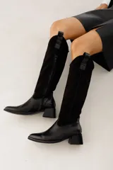 Botas bucaneras negras de cuero y gamuza, con taco de 5cm, punta cuadrada y caña alta. Cuentan con un tirador en la parte superior de la caña.