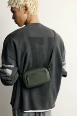 Bandolera porta teléfono de acabado engomado en color verde militar. Cuenta con un compartimento principal y un bolsillo frontal con cierre de cremallera, además de un bolsillo interior también con cierre. Incluye asa de hombro ajustable y extraíble, y detalle de mosquetón en el lateral.