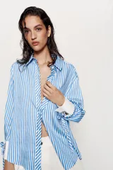 Camisa de cuello solapa y manga larga con rayas verticales celestes y blancas. Cuenta con puños anchos blancos contrastantes, ajustables con hebilla dorada y lazos, y cierre frontal con botones dorados.