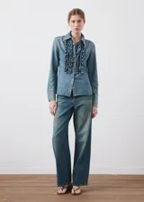 Camisa de denim estilo vaquero, con cuello camisero, manga larga con puños abotonados, cierre frontal con botones y detalle de volantes en el frente.