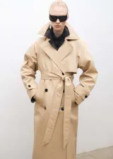 Abrigo tipo trench color beige, de corte oversize y largo hasta la rodilla. Presenta cuello con solapa, cierre frontal cruzado con doble botonadura, cinturón desmontable, dos bolsillos laterales y mangas largas con puños abotonados.