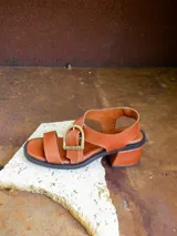 Sandalias de cuero marrón con tiras anchas cruzadas y hebilla metálica decorativa en forma de estribo. Poseen taco cuadrado de altura media y suela de goma negra.