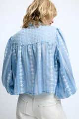Blusa corta de color blanco con diseño de bordados perforados, cuello solapa, cierre frontal con botones y mangas largas abullonadas.