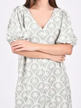 Vestido corto color verde claro con estampado de palmeras blancas en forma de abanico. Tiene escote en V con botones frontales y mangas cortas abullonadas.