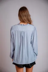 Camisa de color celeste con volados decorativos en el cuello y la parte delantera, cuenta con cordones para ajustar el escote, mangas largas con puños y corte holgado.