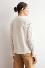 Camisa blanca de manga larga con cuello alto y volados, confeccionada en tela con bordado inglés y detalles calados verticales. Presenta puños con volados y cierre frontal con botones.