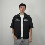 Camisa negra de corte oversize, manga corta y cuello tipo bowling. Presenta botones frontales y estampado con el logo de la marca en el pecho.