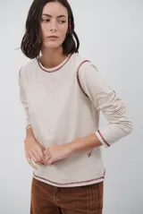 Remera de manga larga en color beige, con cuello redondo y costuras a la vista en color bordó que resaltan el diseño. Presenta un pequeño bordado distintivo en la parte inferior frontal.
