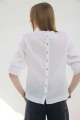 Blusa de lino color marrón, de corte recto y mangas 3/4. Presenta escote redondo con pequeños volados y pliegues frontales, además de detalles de encaje en las mangas y puños con volados.