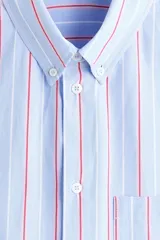 Camisa de corte relajado confeccionada en algodón, con diseño de rayas verticales en tonos azul, blanco y rojo. Presenta cuello americano, cierre frontal con botones, bolsillo en el pecho y mangas largas con puños abotonados.