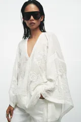 Blusa tipo capa de algodón con escote en pico y mangas largas amplias. Presenta bordados florales a tono en toda la prenda, bajo asimétrico y cierre frontal con botones.