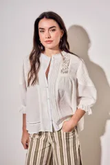 Blusa de algodón peinado color off-white, con cuello mao, cierre frontal de botones, mangas tres cuartos abullonadas con puños con volados, detalles de alforzas y apliques de encaje en el frente y los hombros.