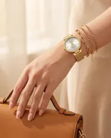 Reloj analógico de mujer con caja redonda dorada, bisel con incrustaciones de cristales y malla de acero tipo milanesa. Esfera minimalista con índices puntuales.