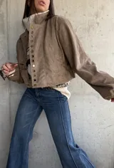 Campera bomber de gamuza color beige, con cierre frontal y detalle de ojales metálicos decorativos en la solapa. Presenta mangas largas con puños elásticos y un corte holgado tipo oversize.