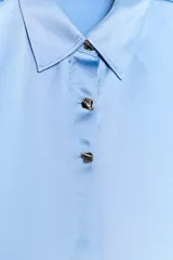 Camisa de cuello solapa y manga larga, confeccionada en tejido satinado con acabado brillante. Presenta cierre frontal con botones y bajo asimétrico.