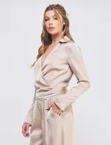 Blusa cruzada color beige de satén, con cuello con solapa, escote en V y lazo de ajuste lateral. Manga larga con pretina, ojal y botón.