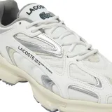 Championes Lacoste modelo L003 2K24, de diseño deportivo y urbano. Presentan una combinación de malla técnica y superposiciones sintéticas en tonos blanco y crema, con detalles plateados. Cuentan con una suela robusta con relieve dentado para mayor tracción y el logo de la marca estampado en el lateral.