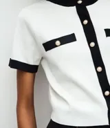 Cardigan de punto de manga corta con cuello redondo y cierre frontal de botones perolados. Presenta un diseño bicolor en blanco y negro con detalles de contraste en cuello, puños, tapetas de bolsillos frontales y vista central.