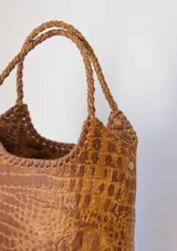 Bolso tipo matera de cuero con textura de reptil, color tostado, con manijas trenzadas y detalles de costura artesanal en los bordes.