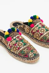 Zueco de punta cerrada con diseño multicolor, decorado con bordados étnicos, lentejuelas, canutillos y pompones de colores. Presenta una suela de yute tipo alpargata con un pequeño taco de madera de 1,5 cm y forro de cuero caprino.