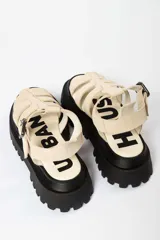 Sandalia tipo gladiadora con plataforma track negra. Presenta un diseño bicolor con tiras delanteras en color amarillo neón y talón con tira ajustable en color beige. Incluye hebilla metálica negra.