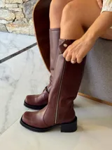 Bota de caña alta estilo biker, confeccionada en cuero color chocolate. Presenta un cierre metálico lateral que recorre toda la caña, estribo y cadena decorativa desmontables, y taco cuadrado con plataforma.
