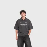 Remera polo gris oscuro de manga corta, con cuello y solapa corta con botones ocultos. Presenta estampado gráfico frontal en color blanco con inscripciones de estilo urbano.