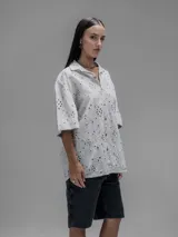 Camisa de manga corta color beige con cuello clásico y cierre de botones. Presenta un diseño de bordado calado con motivos florales y geométricos.