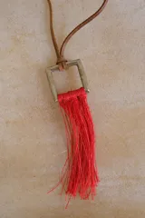 Collar con cordón de cuero tostado y dije geométrico metálico de sección cuadrada con flecos colgantes de hilo rojo.