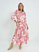 Vestido camisero midi de algodón con estampado floral en tonos rosados y blancos. Tiene cuello camisero, mangas 3/4, botones frontales, falda escalonada y cinturón fino.